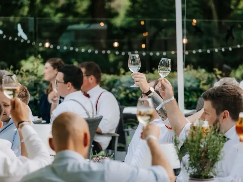 Feiergäste stoßen mit ihren Gläsern an gedeckten Tischen im Biergarten des Haus Pöpsel an.